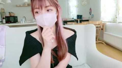 Video of an East Asian woman with straight, light brown hair and pale skin, wearing a black off-shoulder top and a white face mask, sitting on a white sofa in a modern, bright living room.
