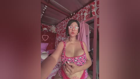 A video of a voluptuous woman with short dark hair, large glasses, and a pink leopard-print top, standing in a red and white patterned room with a bed and stuffed animals.