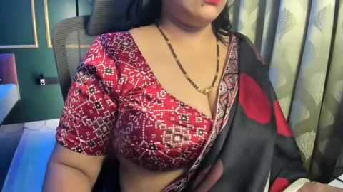 Video of a South Asian woman with medium skin tone, wearing a red and black patterned blouse, black sari with red dots, gold necklace, seated indoors with green and patterned curtains, dark hair.