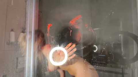 Video of a nude woman with long blonde hair, standing in a shower, water droplets on the glass, illuminated by a circular ring light, reflecting red and orange hues on the glass.