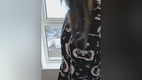 Video of a person with long, dark hair, wearing a black robe with a white cartoon character pattern, standing by a window with a view of an urban landscape.