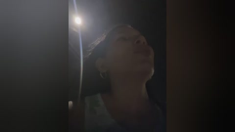 A dimly lit video of a woman with short brown hair, wearing a dark hat, gazing upward with a serene expression, against a dark background.