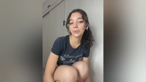 Video of a young woman with curly dark hair, wearing a black t-shirt, sitting against a white wall in a minimalistic room with an air conditioning unit.