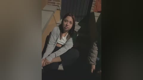 A video shows a young woman with straight brown hair, wearing a gray hoodie and black pants, sitting on a staircase, leaning against a wall.