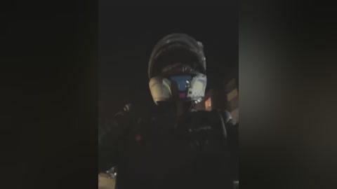 Video of a person wearing a dark, hooded jacket with a reflective visor, standing in a dimly lit, dark room, with indistinct background, conveying a sense of mystery and caution.