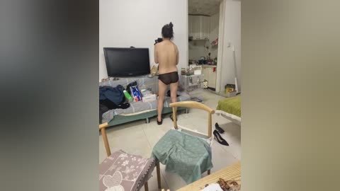 A video captures a topless woman with a small build, wearing only black panties, standing in a messy living room. The room is cluttered with clothes, a TV, and a bed.