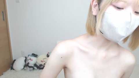 A video of a topless, fair-skinned woman with short blonde hair wearing a white mask, standing in a minimalist room with a fluffy white carpet and a stuffed panda bear on the floor.