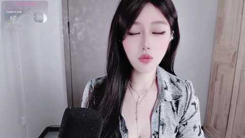 Video of a young Asian woman with long black hair, wearing a patterned shirt, and a gold necklace, standing in a bathroom with a shower curtain and wooden door in the background.