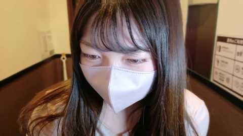 Video of an Asian woman with straight, dark hair and bangs, wearing a white face mask, light shirt, in a dimly lit hallway with beige walls and a wooden floor.