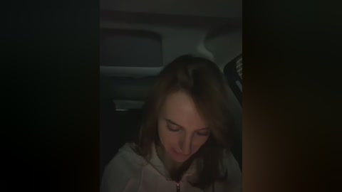 Video of a young woman with shoulder-length brown hair, wearing a light-colored jacket, sitting in a dark car interior, looking down with a sad expression.