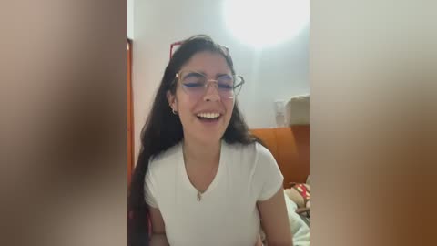 Video of a young woman with long, dark hair, wearing glasses, smiling in a dimly lit room with white walls and a bed.