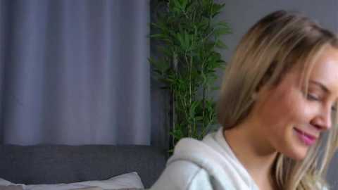 Video of a fair-skinned woman with blonde hair, wearing a white hoodie, smiling softly, standing in a dimly lit room with a grey curtain and a potted bamboo plant in the background.