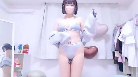 Video of an Asian woman in a white bra and panties, with a mask, lifting a white jacket in a cluttered, brightly-lit room with clothes hanging on hooks and a box of tissues on a shelf.