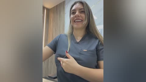 Video of a smiling Latina woman with long, straight hair, wearing a dark blue scrub top. She stands indoors, possibly in a medical setting, with beige curtains and a tiled wall in the background.