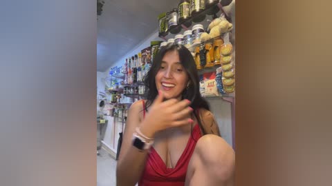 Video of a smiling Latina woman with long black hair, wearing a red dress, sitting in a messy kitchen with shelves full of condiments and utensils.