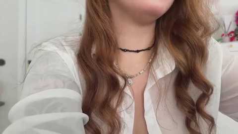 Video of a fair-skinned woman with long, wavy brown hair, wearing a sheer white blouse, a black choker necklace, and a delicate silver chain necklace. Background features a blurred, white interior with a hint of red and blue decor.
