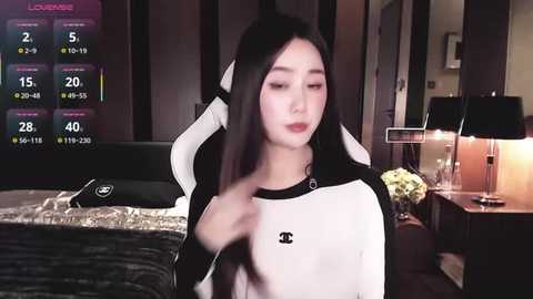 Video of a young East Asian woman with long black hair, wearing a black-and-white Chanel hoodie, seated in a dimly lit bedroom with a gaming setup.