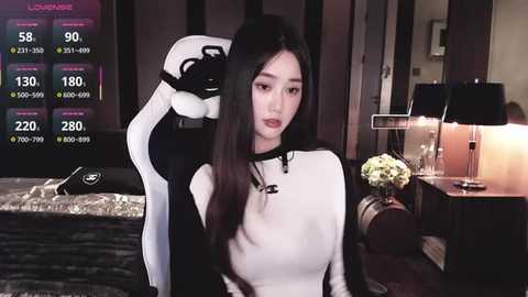 Video of an East Asian woman with long black hair, wearing a black and white sweater, sitting in a gaming chair, in a dimly lit room with a table lamp and flowers.