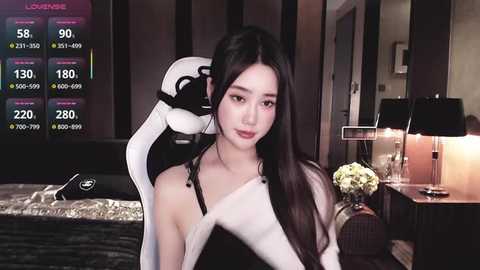 A video shows an Asian woman with long black hair, wearing a black dress and headphones, in a modern bedroom with a bed, lamp, and flowers.