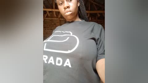 Video of a young African woman with medium brown skin, wearing a black t-shirt with a white \"RADA\" logo, and long braided hair. Background shows a rustic wooden ceiling.