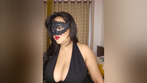 A video of a woman with long black hair, fair skin, wearing a black lace mask, revealing her large breasts in a low-cut black dress. The background shows curtains and a bed, suggesting an indoor setting.