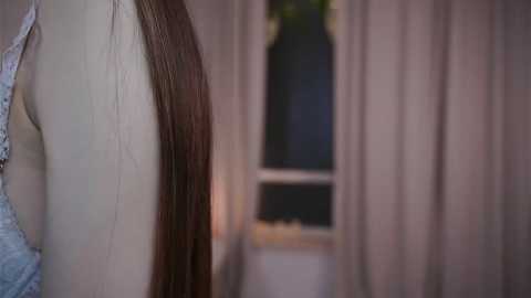 Video of a woman with long, straight brown hair, wearing a lace bra, standing next to a tall, narrow window with closed, beige curtains. The background is soft and blurred, enhancing the focus on her hair and bra.