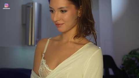 Video of a fair-skinned, brunette woman in a white, off-the-shoulder top, revealing a lace bra, indoors with blurred background.