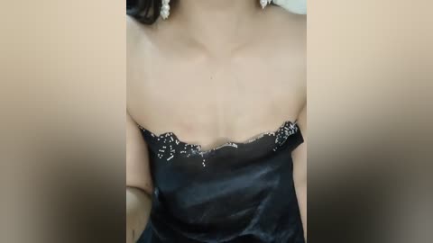 A close-up video of a woman wearing a black, satin strapless dress with delicate white lace trim, revealing her bare shoulders. She has light skin and is indoors against a beige background.