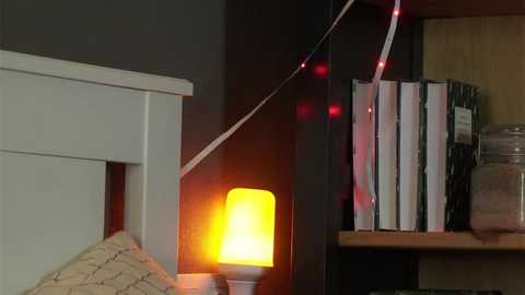 Video of a cozy bedroom with a white headboard, a warm, glowing lamp, a bookshelf with books and boxes, and red string lights draped on a dark wall.