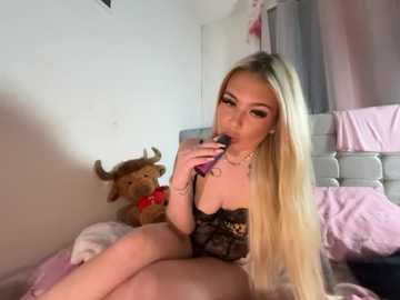 Video of a blonde woman with long hair, wearing black lace lingerie, holding a purple vibrator, sitting on a bed with a stuffed reindeer and a white tufted headboard.