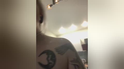 Video of a person's back in a dimly lit room, showing a tattoo of a crescent moon and a small bird. The background is blurred, featuring a ceiling light and a desk.