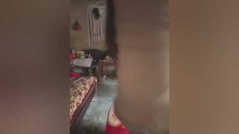 A video captures a dimly lit, cluttered bedroom with a person's lower body, a bed with patterned sheets, and a desk with scattered items in the background.