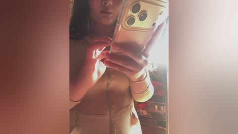 Video of a woman with light skin, medium breasts, and long dark hair, wearing a beige zip-up jumpsuit, taking a selfie with an iPhone in a dimly lit room.