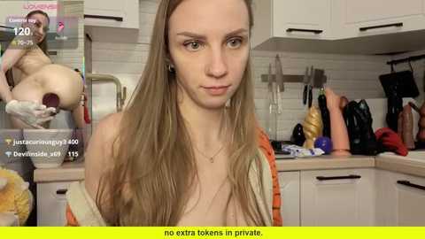 Video of a young, fair-skinned woman with long, straight blonde hair, sitting topless in a modern kitchen, surrounded by various kitchen utensils. The background shows white cabinets and a tiled backsplash.