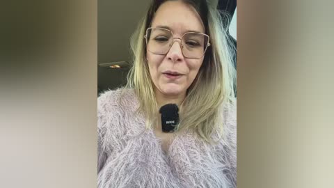 Video of a middle-aged Caucasian woman with blonde hair and glasses, wearing a fluffy pink sweater and a black medical alert bracelet, indoors with blurred background.