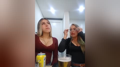 Video of two women with fair skin, long blonde hair, and large breasts, in a modern, brightly lit room. One woman in a maroon top, the other in a black top, adjusting her glasses.