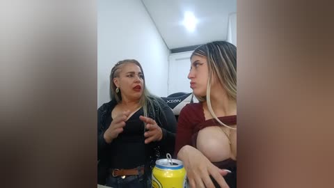 A candid video shows two women in a dimly-lit room. The woman on the left has long blonde hair, red lipstick, and a black top, gesturing. The woman on the right, with long blonde hair, a maroon top, and large breasts, holds a can of soda.