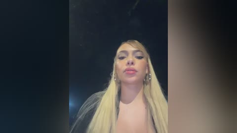 Media: Video of Miley Cyrus, a young Caucasian woman with long, platinum blonde hair, wearing a black top and large hoop earrings. She has a fair complexion and full lips. The background is dark, making her stand out.