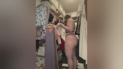 Video of a nude, curvy, light-skinned woman with auburn hair, wearing only a brown thong, standing in a cluttered closet with hanging clothes and storage bins, in a dimly lit room.