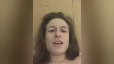 A video of a young Caucasian woman with shoulder-length brown hair, wearing a light-colored top, captured in a bathroom with wooden walls and a mirror reflection.