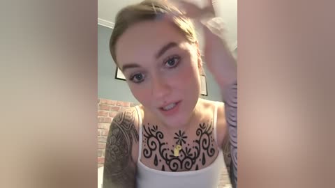 Video of a young woman with light skin and blonde hair, wearing a white tank top, showcasing intricate black floral tattoos on her chest and arms, smiling in a bathroom with a brick wall and soft lighting.