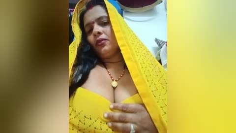 A video shows a South Asian woman with medium skin tone, wearing a bright yellow saree and a matching blouse, revealing her ample cleavage. She has long, dark hair and a neutral expression. The background is indistinct.