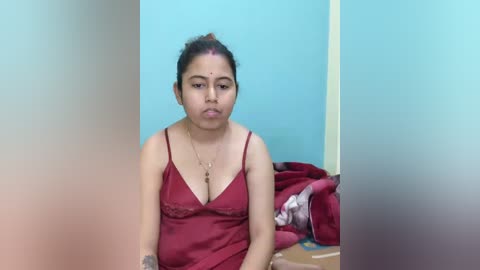 A video of a South Asian woman with medium-brown skin, dark hair tied back, wearing a red satin camisole, sitting indoors against a turquoise wall, with a bed and clothes visible in the background.
