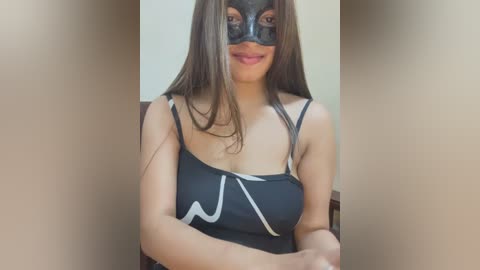 Video of a woman with medium skin tone, long brown hair, wearing a black mask with a white pattern and a black spaghetti-strap top. She sits indoors against a beige wall, smiling slightly.