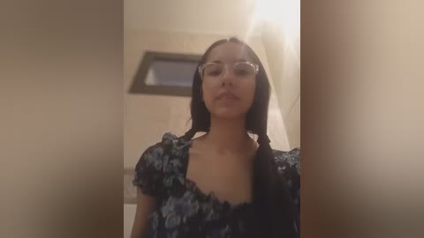 Video of a young woman with straight, black hair parted in the middle, wearing glasses and a floral-patterned dress, standing in a dimly lit room with beige walls and a framed picture in the background.