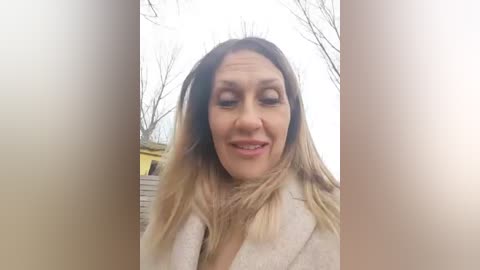 Video of a smiling, light-skinned woman with long, blonde hair, wearing a beige coat, standing outdoors in a suburban neighborhood with leafless trees and a yellow house in the background.