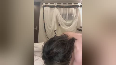 Video of a person with dark hair, seen from behind, lying on a bed with white sheets. A beige curtain with a wooden pole hangs above. The room has a neutral, warm-toned decor.