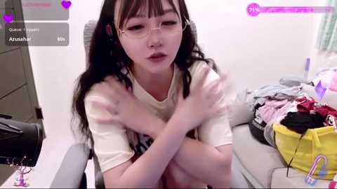 A candid video of a young Asian woman with long black hair, wearing glasses, and a white T-shirt, sitting on a bed with messy clothes and a yellow bag.