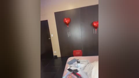 Video of a dimly lit, cluttered bedroom with a bed, red balloons, and a dark wooden wardrobe. The room has a beige wall and dark flooring.