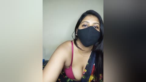 Video of a woman with medium skin tone, long black hair, wearing a black face mask, floral blouse, and pink bra, looking into the camera.